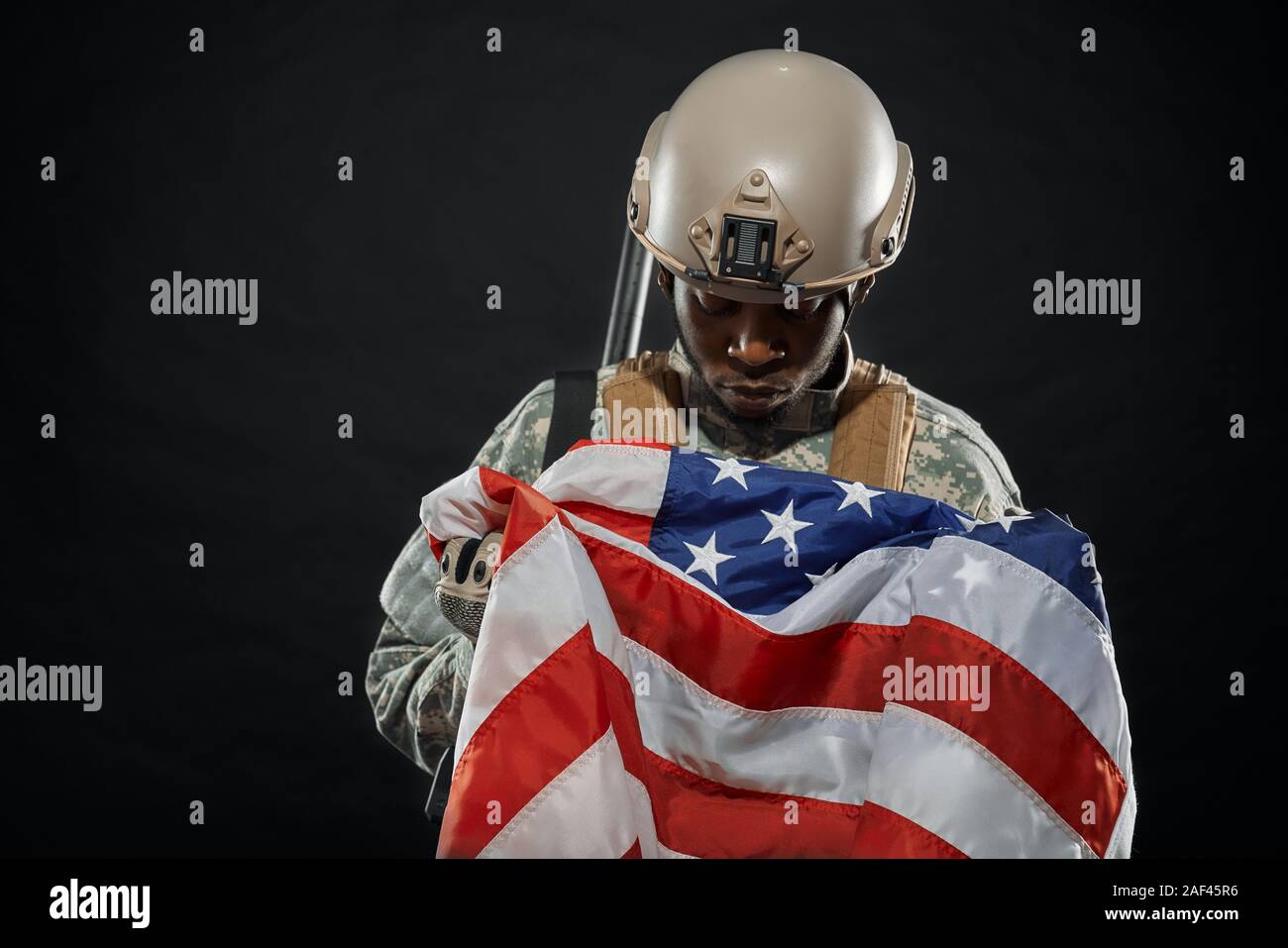 Front view of American soldier wearing uniform and helmet on head holding nationals flag in hands. Ranker sadly looking on American flag. Black isolated background. Stock Photo