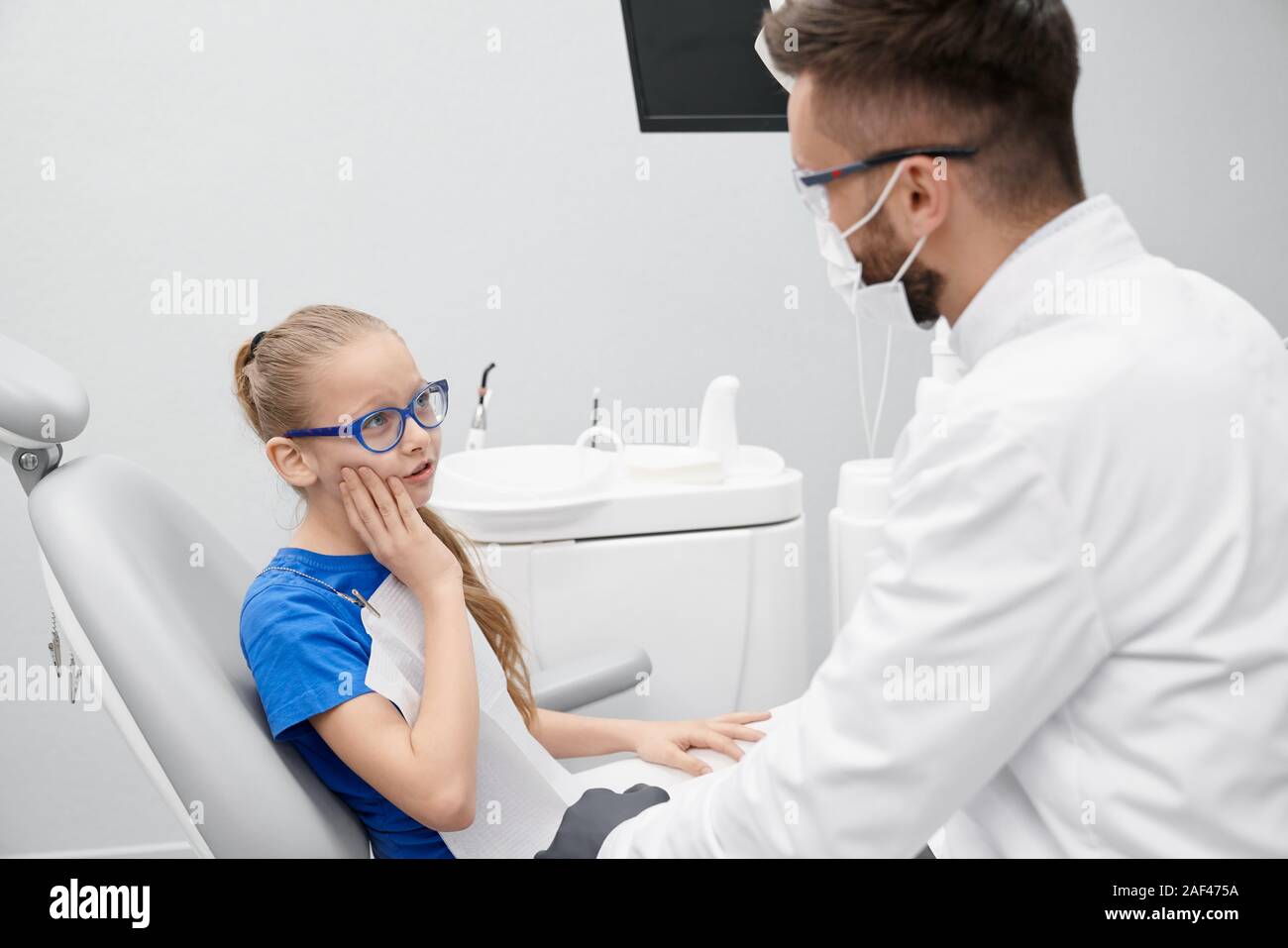 Side view of little patient complaining about pain in teeth in clinic. Girl in glasses sitting in dentist office, looking at doctor and talking. Concept of treatment and stomatology. Stock Photo