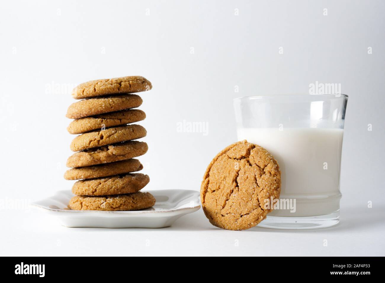 A glass of milk with a ginger cookie leaning against it next to a stack of ginger cookies on a white plate with a white background; copy space Stock Photo