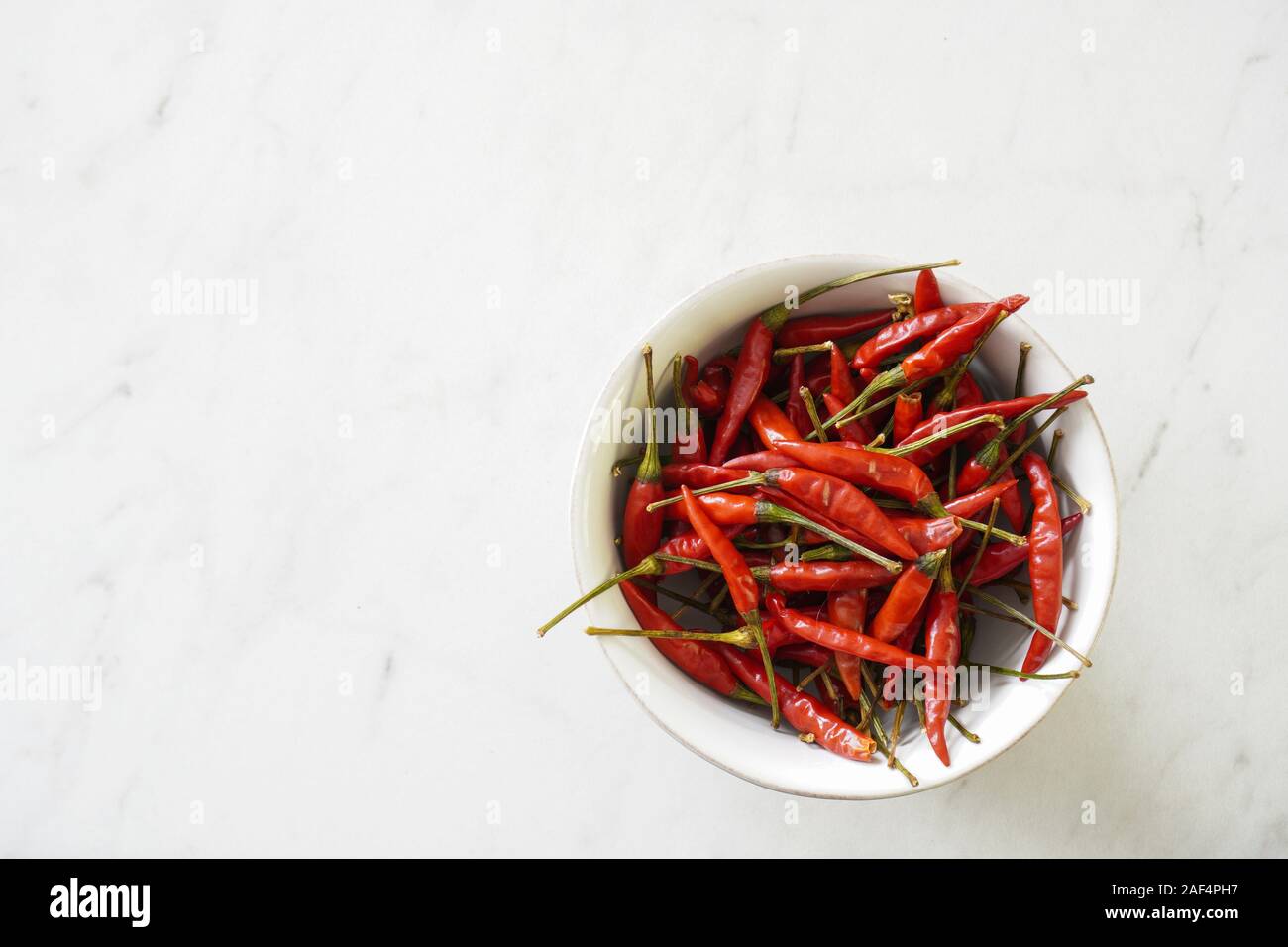 A white bowl of bright red Thai bird chiles on a marble cutting board with copy space; food preparation Stock Photo