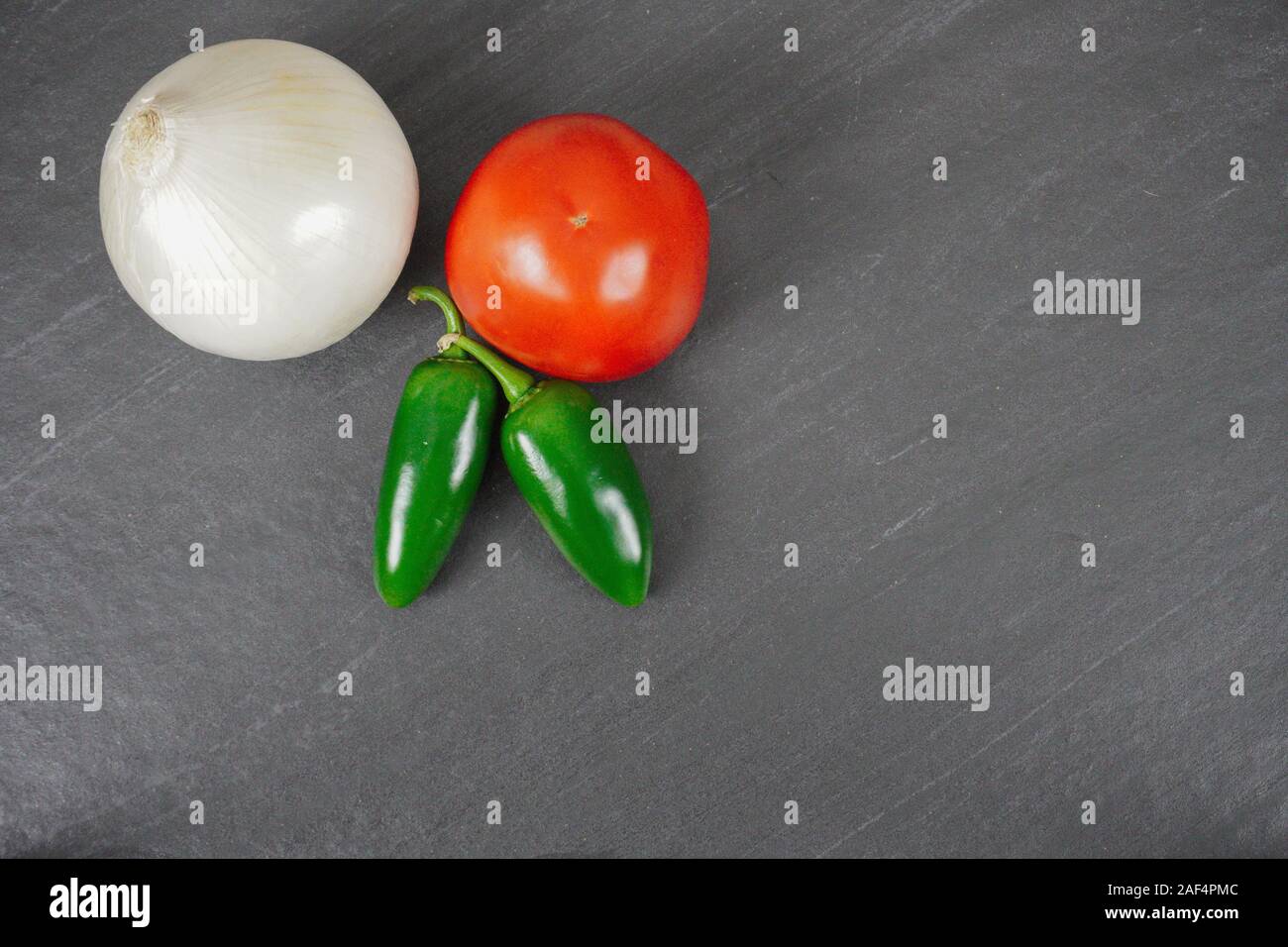 Ingredients for salsa- jalapenos, a tomato and an onion- rest on a gray slate cutting board- food preparation Stock Photo