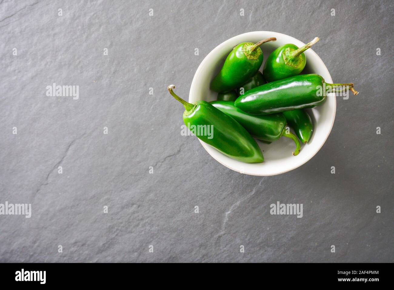 A white bowl filled with bright green jalapeno peppers rests on a gray slate cutting board; food preparation Stock Photo