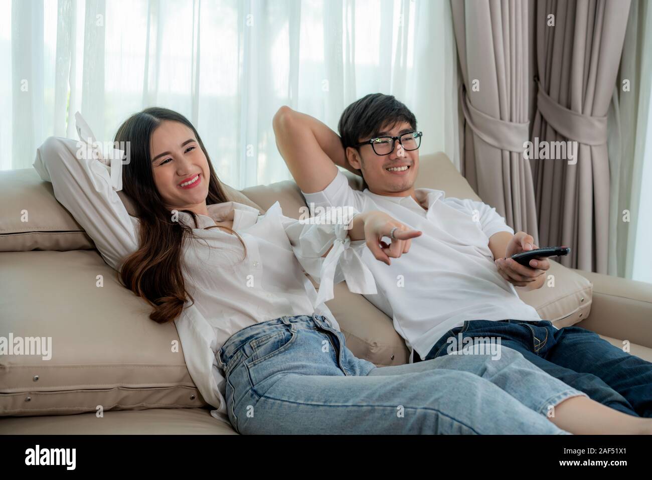 Asian couple lifestyle, man holding remote control and woman watching TV movies at home, family lifestyle relax and recreation concept. Stock Photo