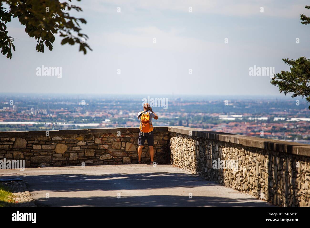 the tourist man stands with his back on the viewing platform in Bergamo in  Italy and takes a photo on the camera wearing shorts, sandals and an orange  Stock Photo - Alamy