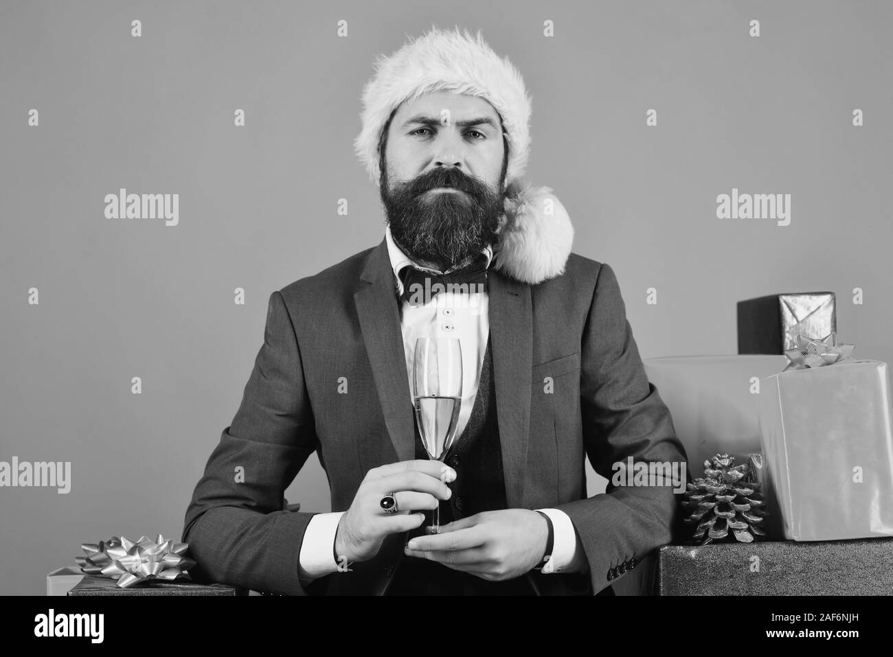 Santa in retro suit near blue and red gifts on red background. Businessman with proud face by stack of boxes and glass of drink. Man with beard holds champagne. New year eve concept. Stock Photo