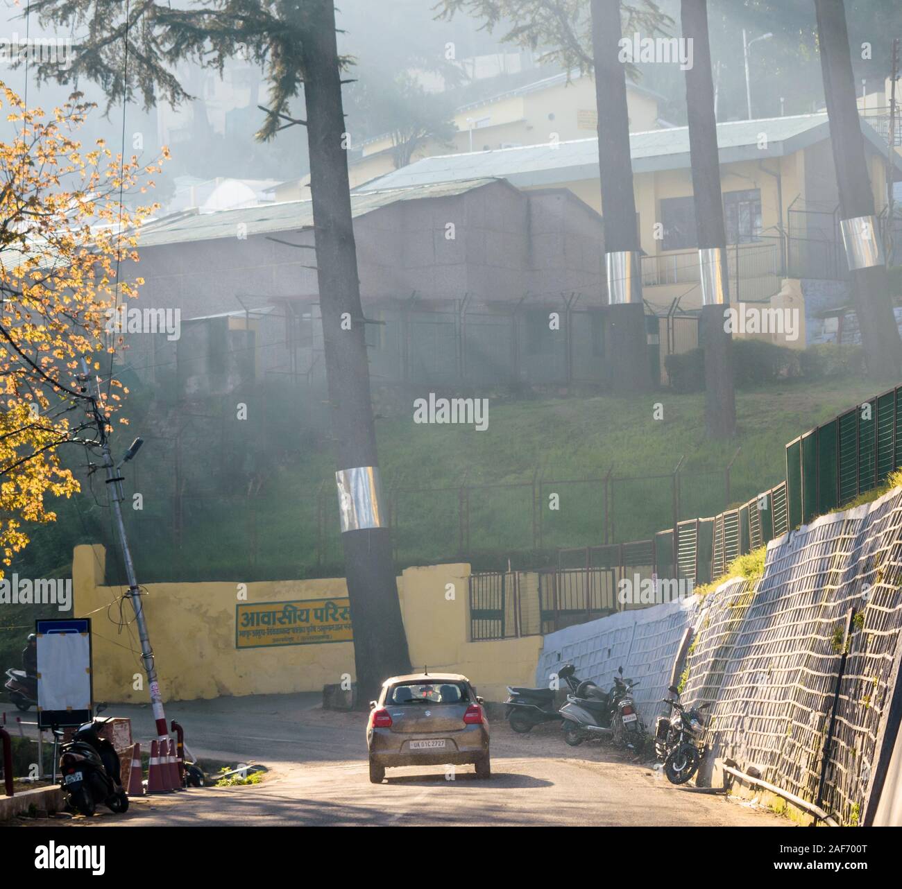 Shafts of light hit the trees and the buildings of the Almora Cantontment in the early morning mist. A Maruti Suzuki drives down the road. Stock Photo