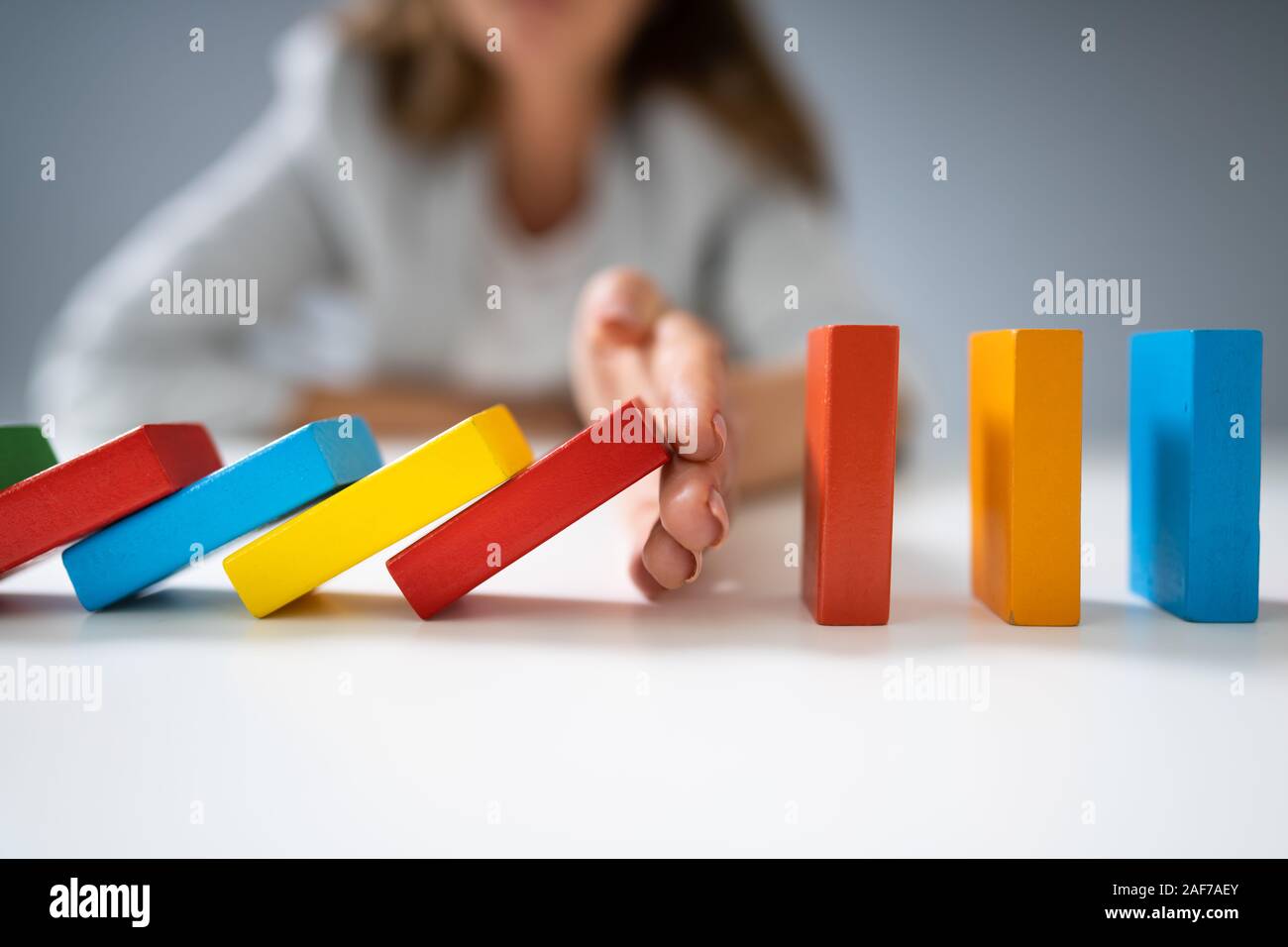 High Angle View Of A Businessperson Stopping Colorful Dominoes From Falling On Desk Stock Photo