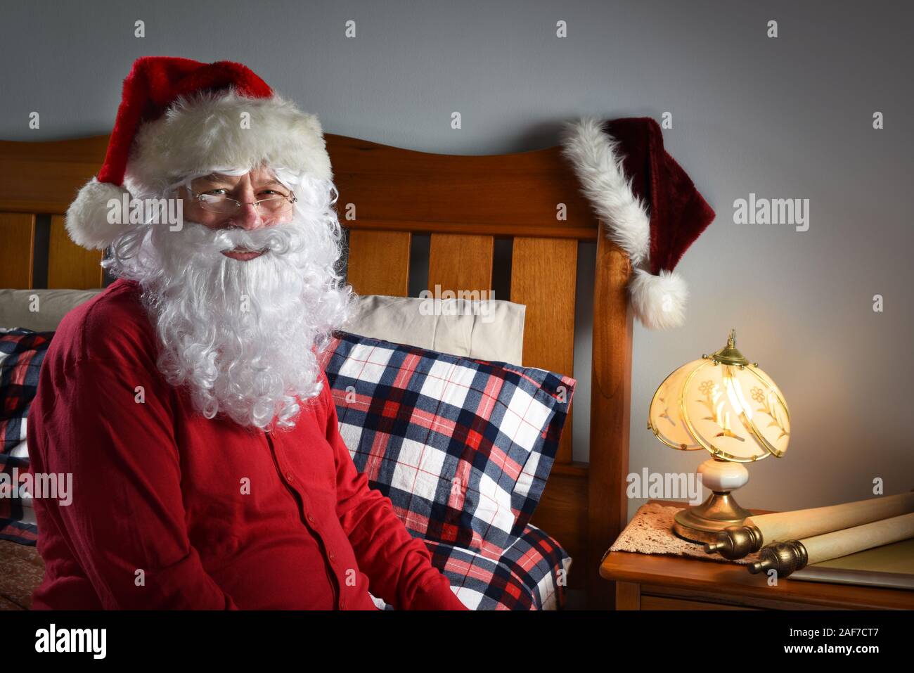 Closeup of Santa Claus wearing a red union suit sitting on the edge of his bed at the North Pole, ready to go to sleep after delivering packages on Ch Stock Photo