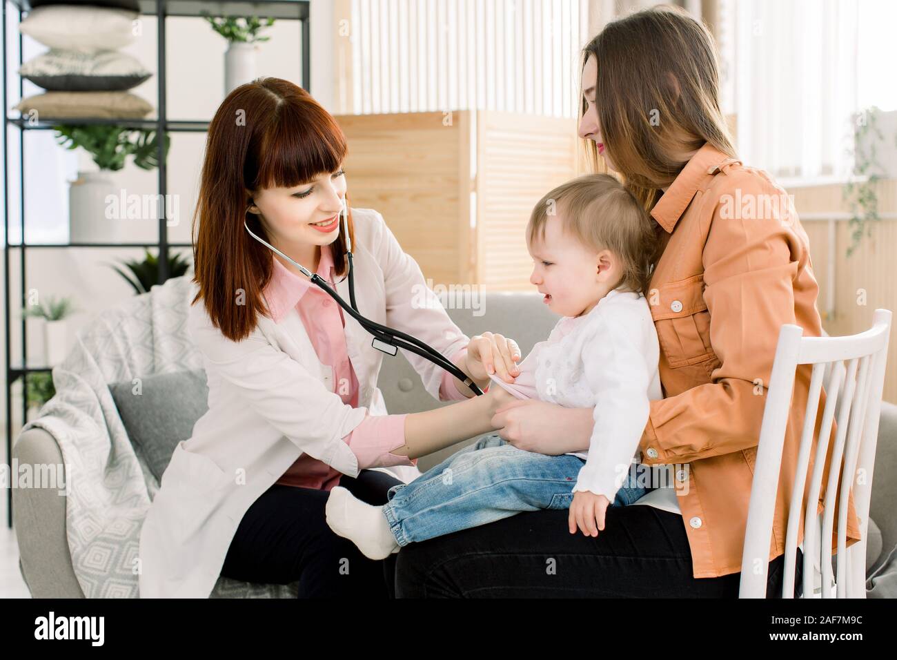 crying sick kid baby girl on mother hands in hospital or at home, and female GP doctor having examination. Stock Photo