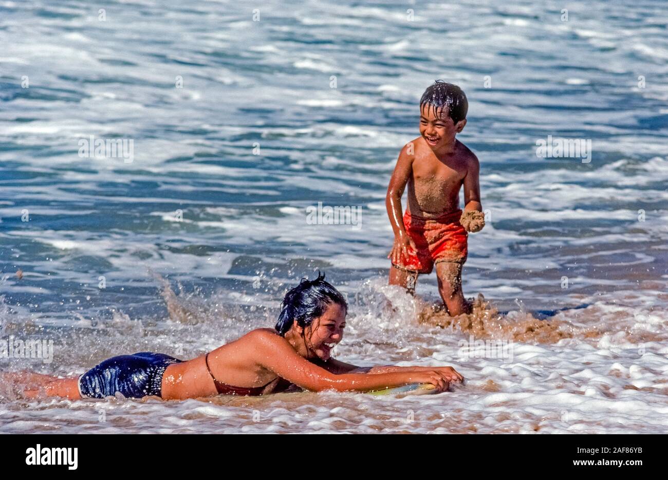 Two Native Hawaiian children play in the sandy Pacific Ocean surf along the shore of Molokai Island in Hawaii, USA. The young boy is ready to throw a handful of wet sand at his sister who is floating in the water on a skimboard. Molokai is the fifth largest of eight major islands in the Aloha State but a lesser known tourist destination. A major attraction for adventurous travelers is a mule ride down steep seaside cliffs to visit the isolated site of a 19th-Century leper colony on Molokai's rugged northern coast. Stock Photo