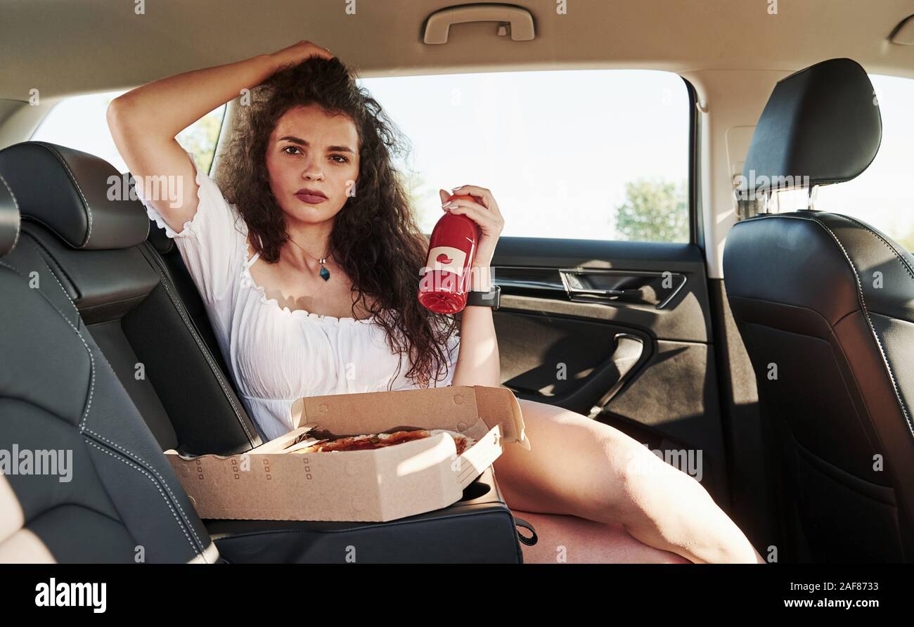 Brunette with curly hair sits in the modern car with bottle with tomato juice in hand and pizzas Stock Photo