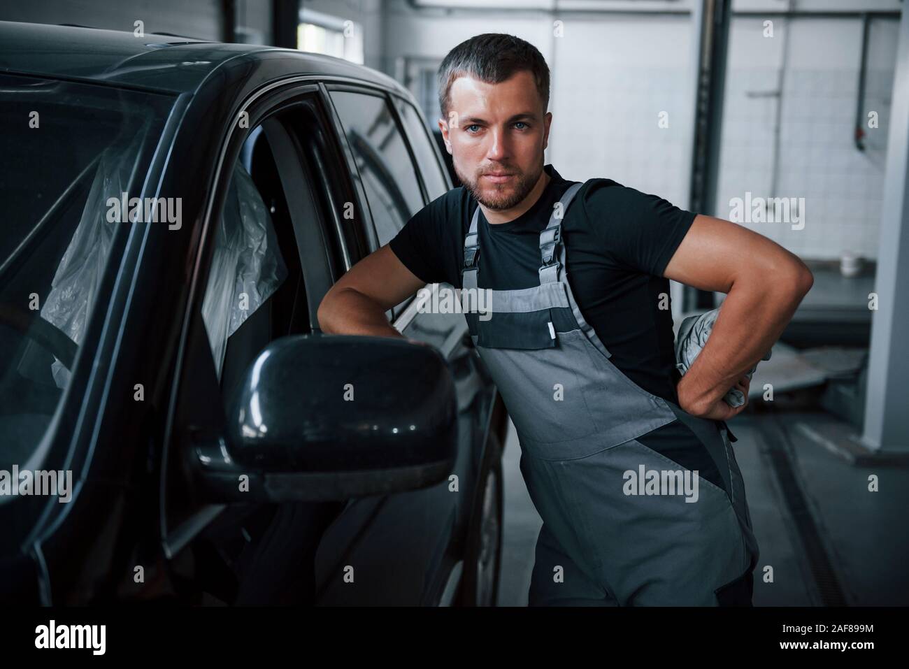 Looks straight at the camera. Young mechanic stands indoors near black
