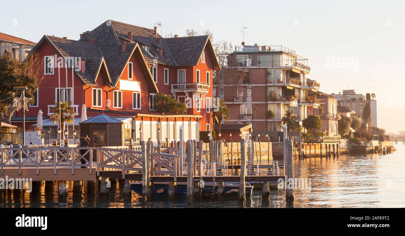 The waterfront of Lido , Venice, Veneto, Italy at sunset with colorful red restaurant  and apartment buildings. Wooden jetty for ferry in foreground. Stock Photo