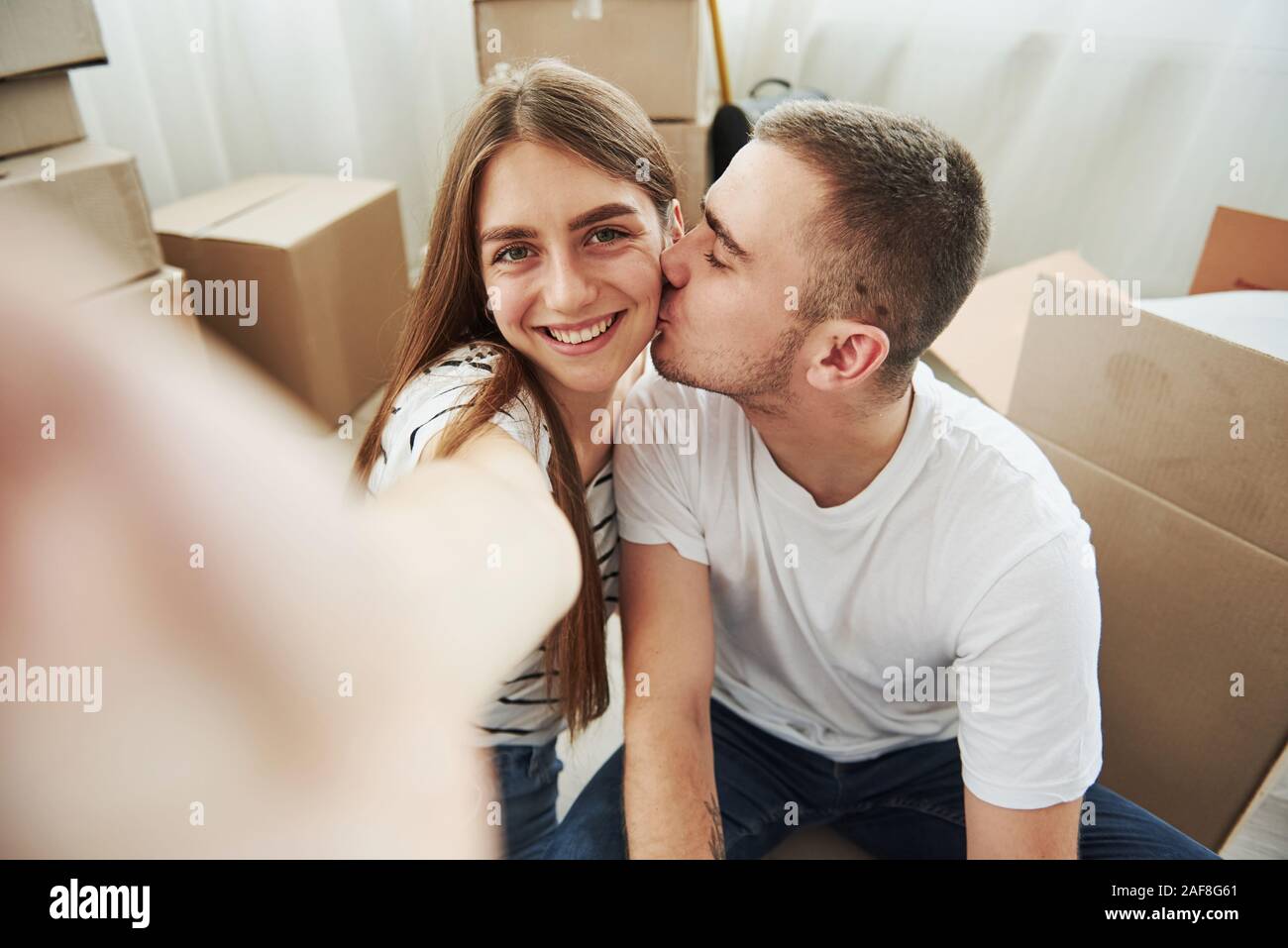 Making a photo. Guy kisses his wife. Cheerful young couple in their new apartment. Conception of moving Stock Photo