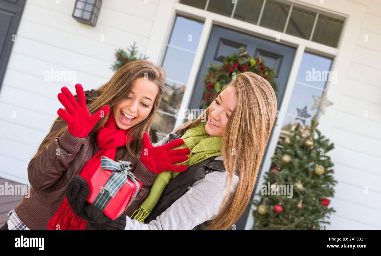 Young Mixed Race Girls Holding Wrapped Gift Standing on Christmas Decorated Front Porch . Stock Photo