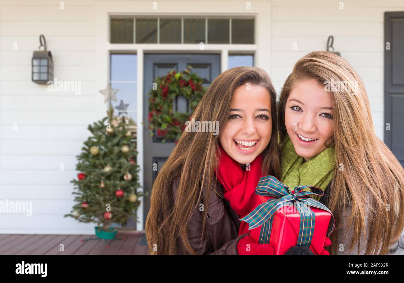 Young Mixed Race Girls Holding Wrapped Gift Standing on Christmas Decorated Front Porch . Stock Photo