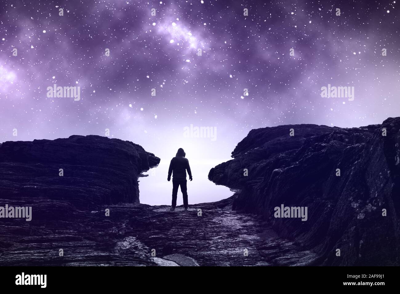 A lone spooky hooded figure, standing on the edge of the sea. With a muted, blue edit. With a beautiful mysterious background of the universe and star Stock Photo