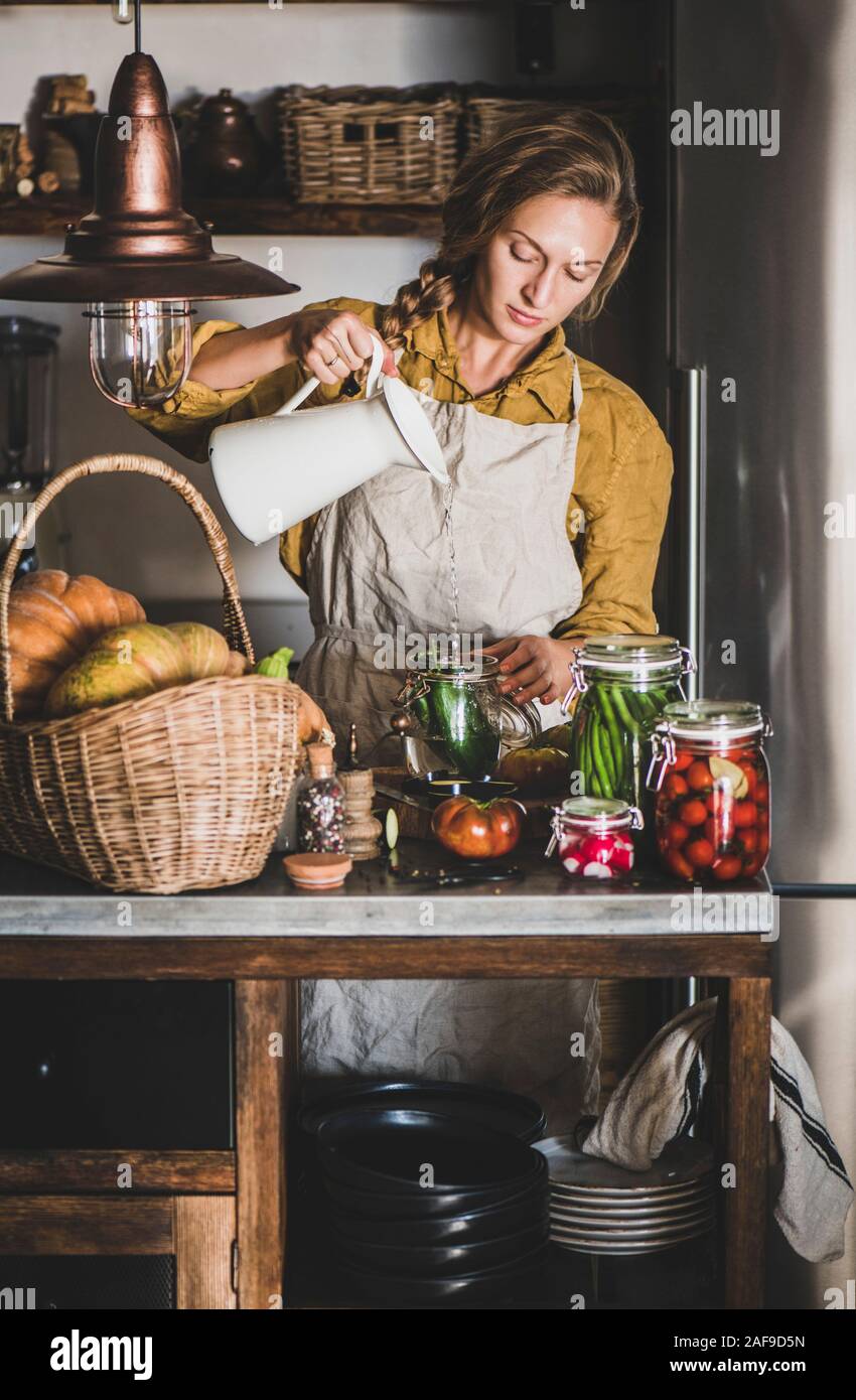 Autumn seasonal vegetable pickling and canning. Young blond woman in linen apron adding water to cucumbers and cooking homemade vegetables preserves i Stock Photo