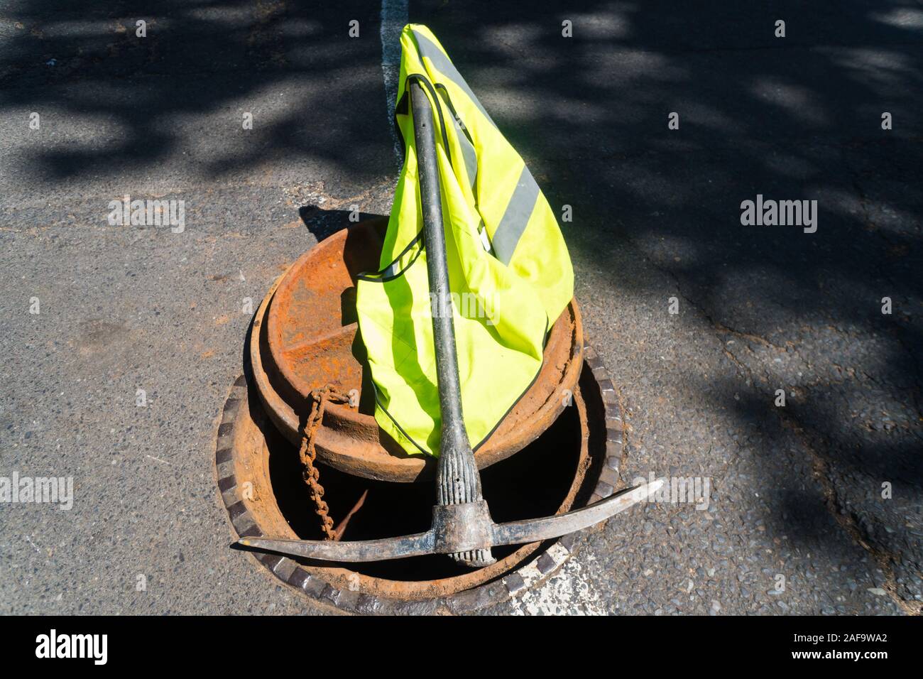 open manhole cover with a pickaxe and high visibility vest resting on it during road works concept blocked drains and sewers and roadworks Stock Photo