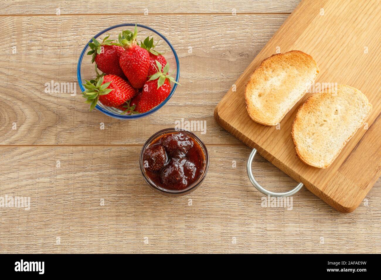Homemade strawberry jam in a glass bowl, fresh berries, toasts on cutting board on wooden desk. Top view. Stock Photo