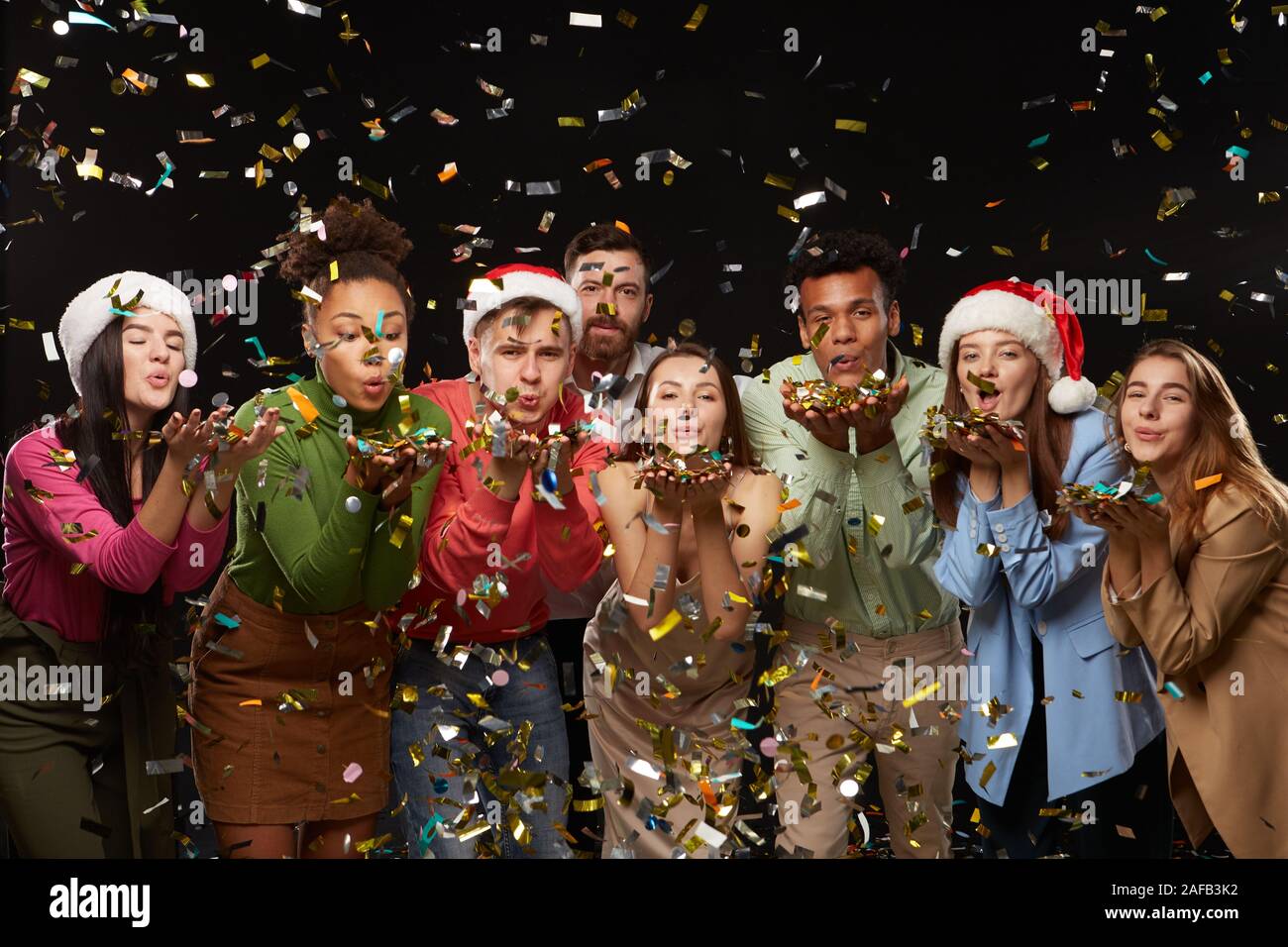 Group of cheerful young people are clinking with champagne glasses. On friends from above the festive confetti falling. Shooting in professional studio on isolated black background. Stock Photo