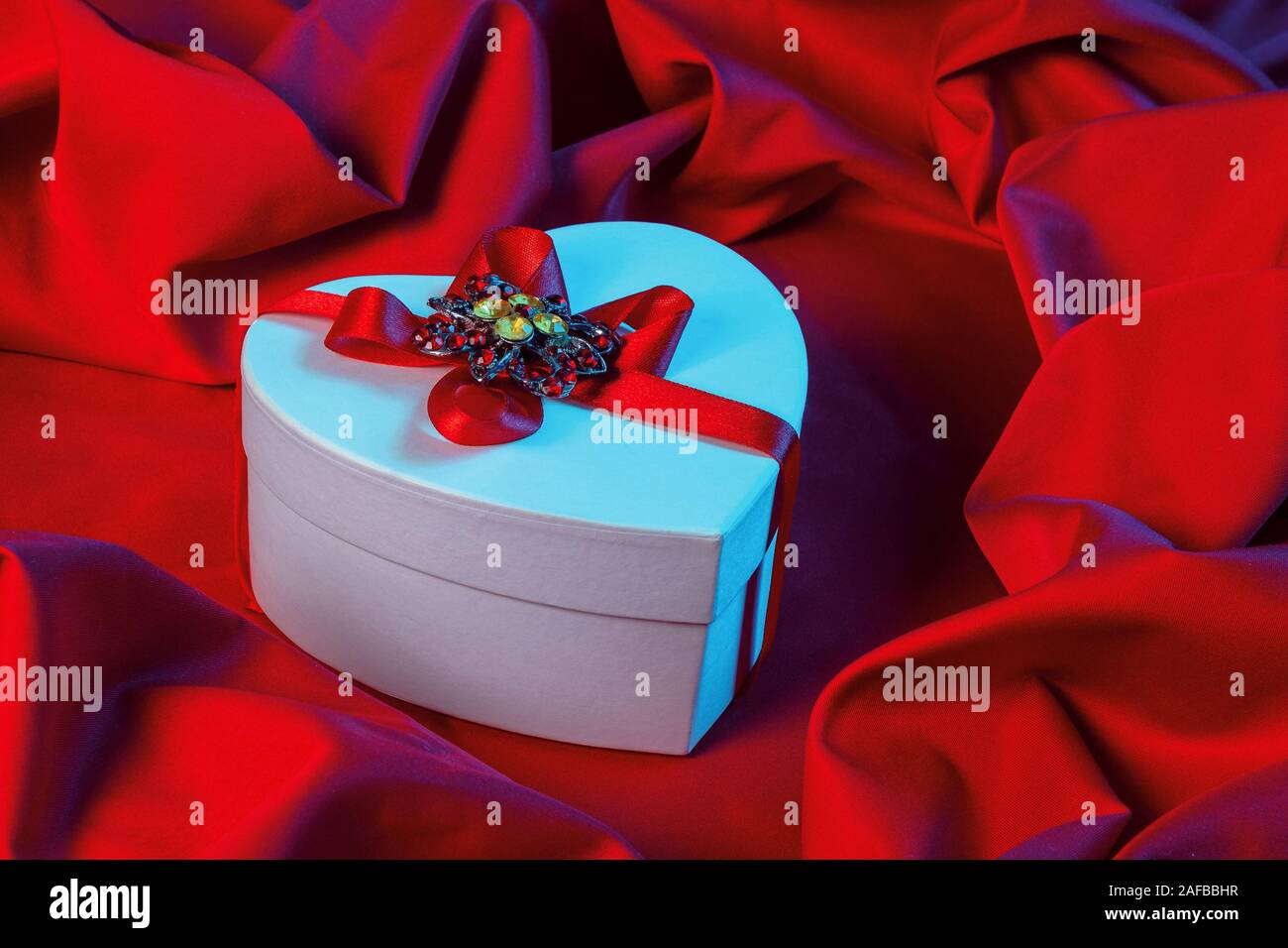 valentines greeting card in blue light. white cardboard box in shape of heart wrapped in ribbon lay on a red cloth which repeats the form of present p Stock Photo