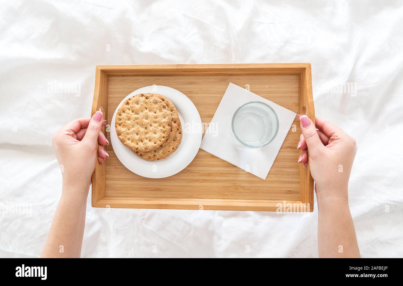 Woman holding tray with diet breakfast of crackers and water on bed