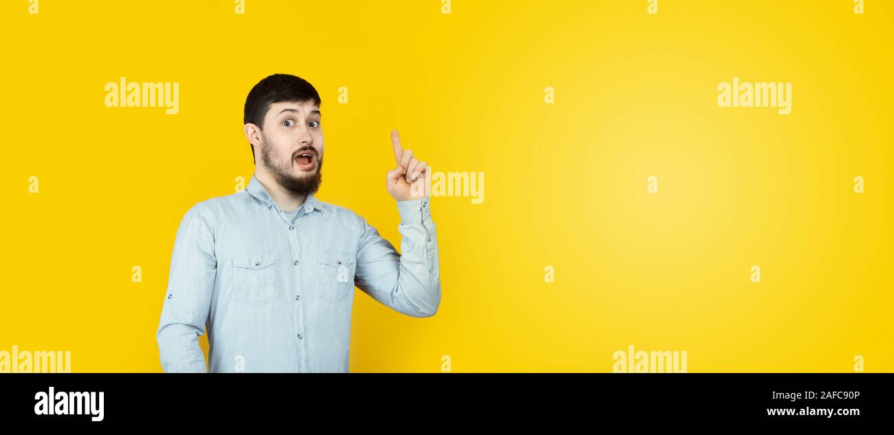 Photo of caucasian man  reliable person got new brilliant idea, startup raise finger, wear jeans denim shirt  over yellow background, panoramic mock-u Stock Photo