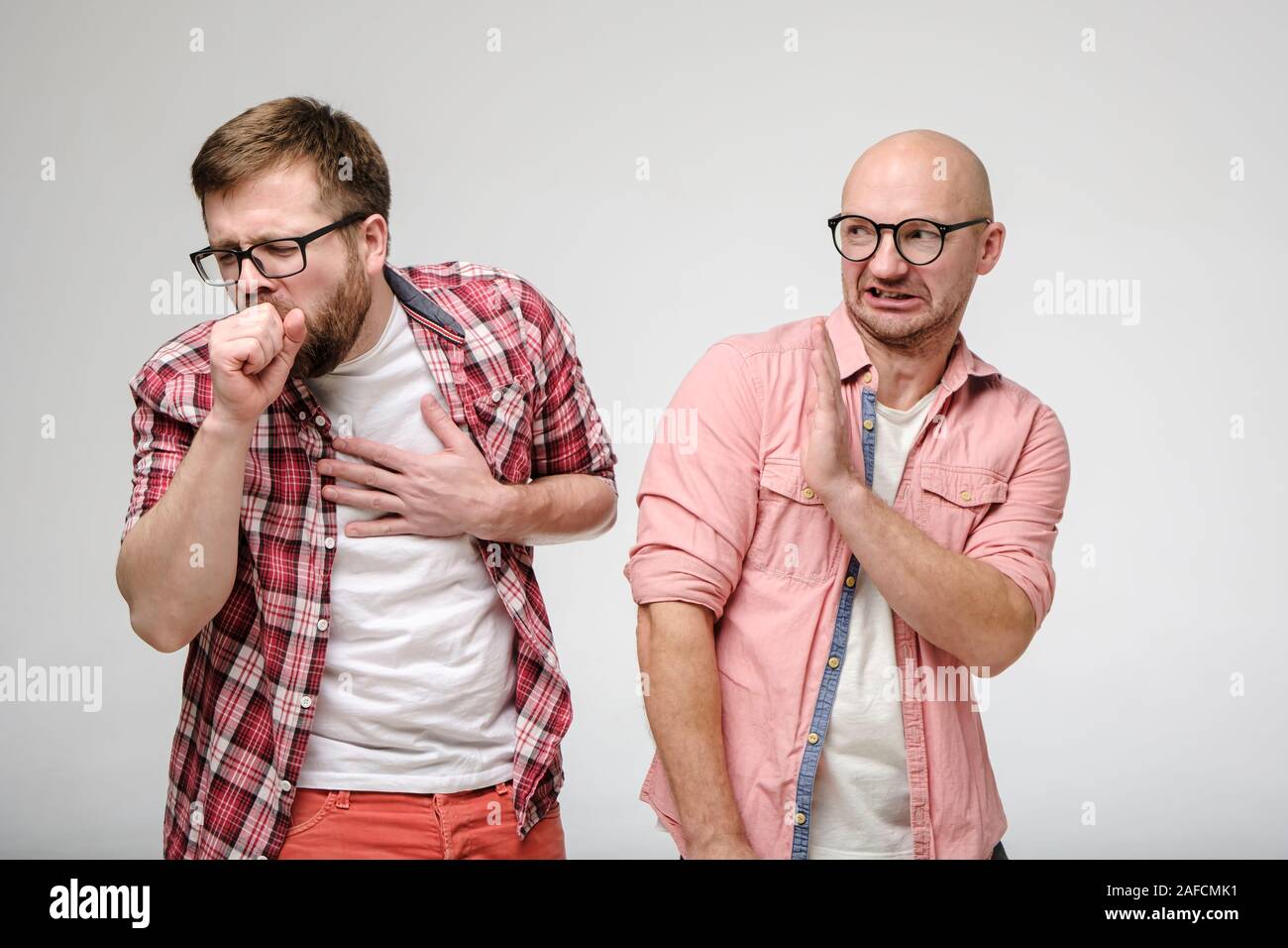 One man coughs, closing mouth with hand, and other man makes a gesture of protection, putting palm forward and looking in disgust. Stock Photo