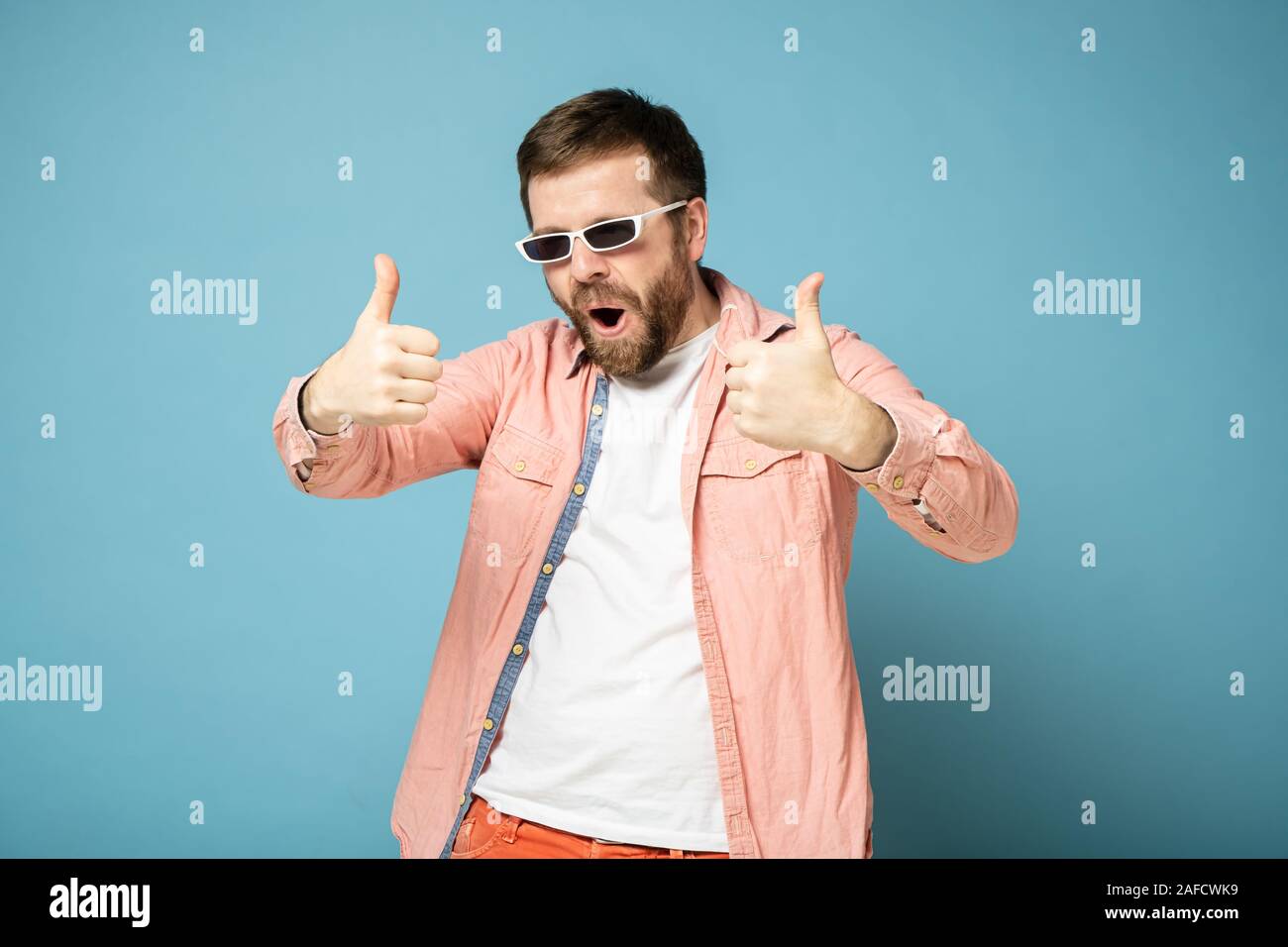 Strange, bearded man in sunglasses gestures show thumbs up on two hands, opens mouth and makes a weird expression on face. Stock Photo