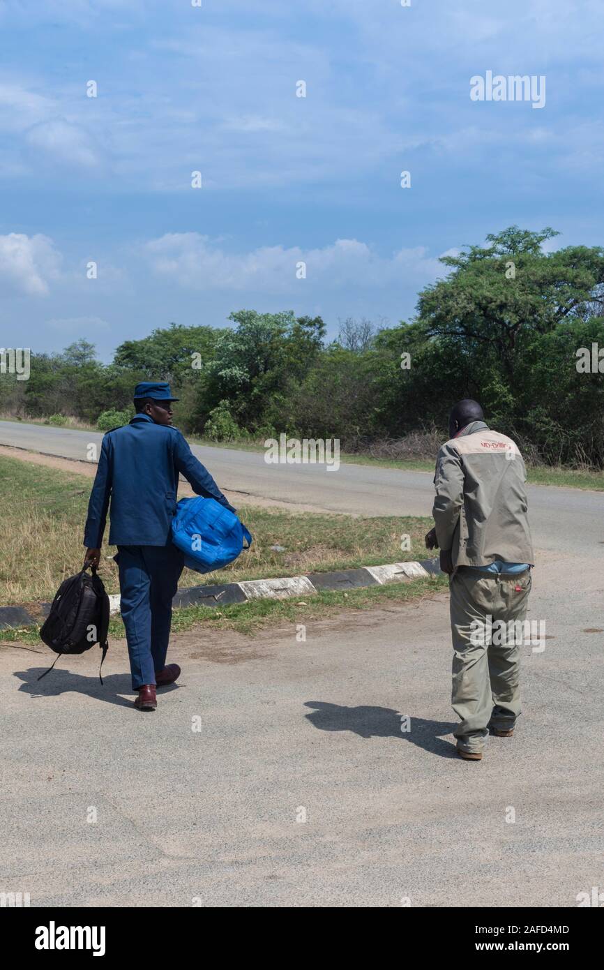 Zimbabwe. A prisoner and a prison guard walk on road, waiting for a ride. Due to the economic situation, the prison service cannot send prison cars everywhere needed, so it's cheaper just to send a warden with the prisoners to hitch a ride or to take public transportion. Stock Photo