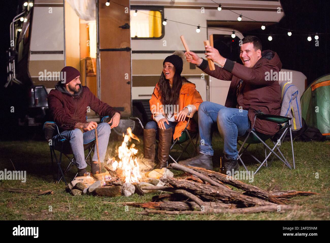 Group of close friends warming up around camp fire and laughing together. Retro camper van. Stock Photo