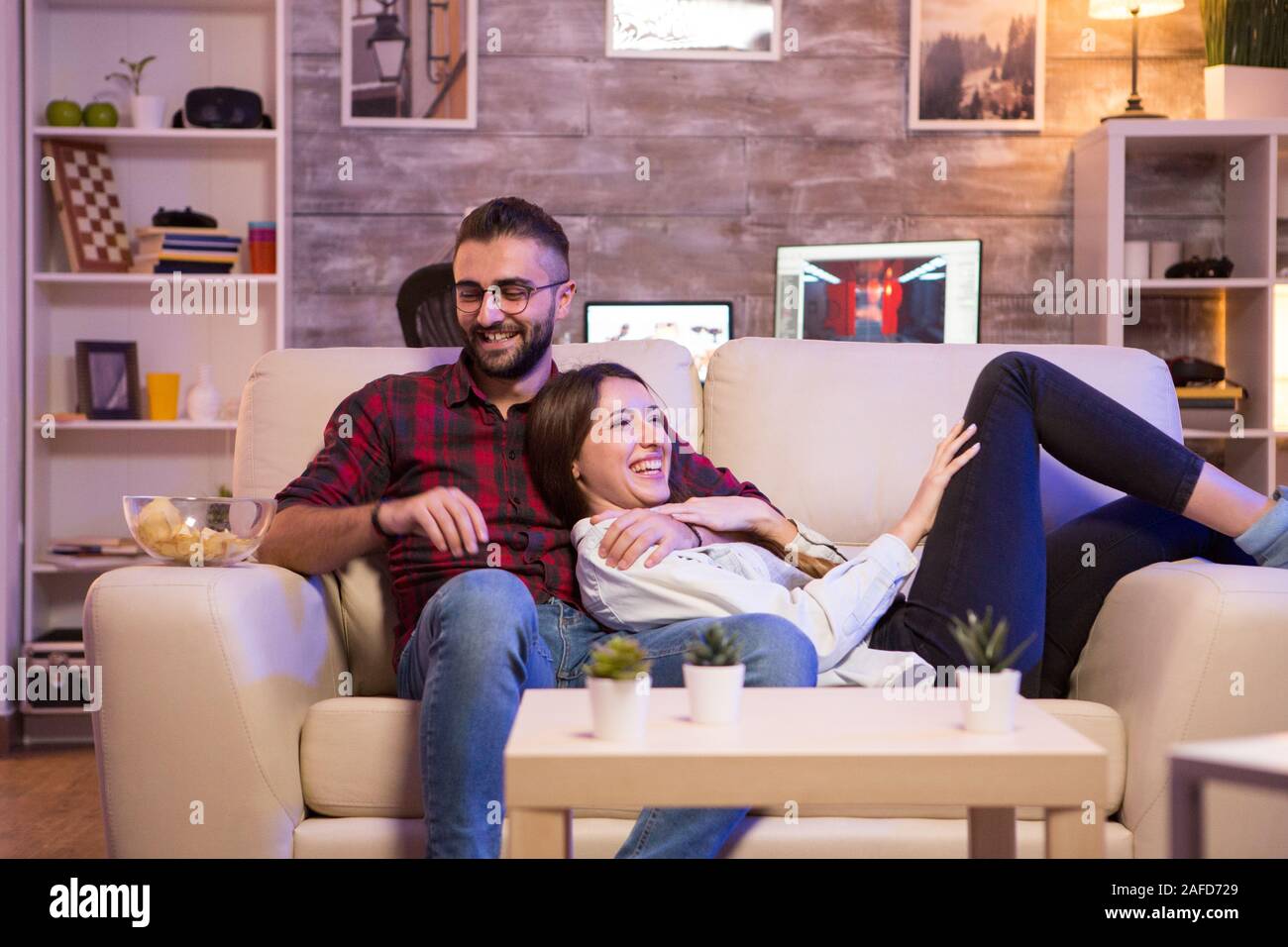 Cheerful young couple laughing while watching a tv show on tv at night. Couple sitting on sofa. Stock Photo