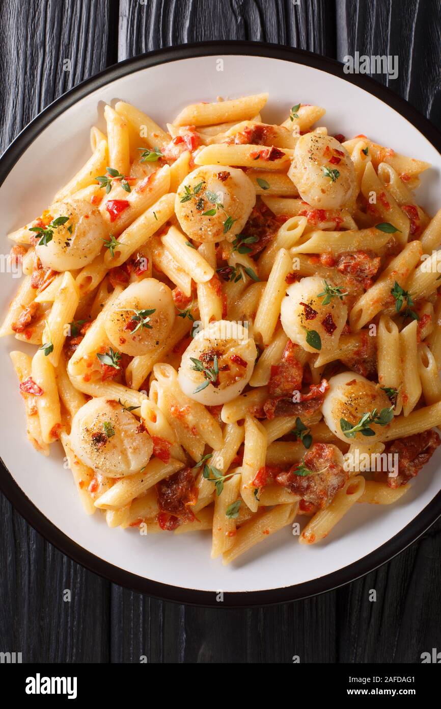 Penne pasta with scallops, tomatoes, herbs and spicy sauce close-up in a plate on the table. Vertical top view from above Stock Photo