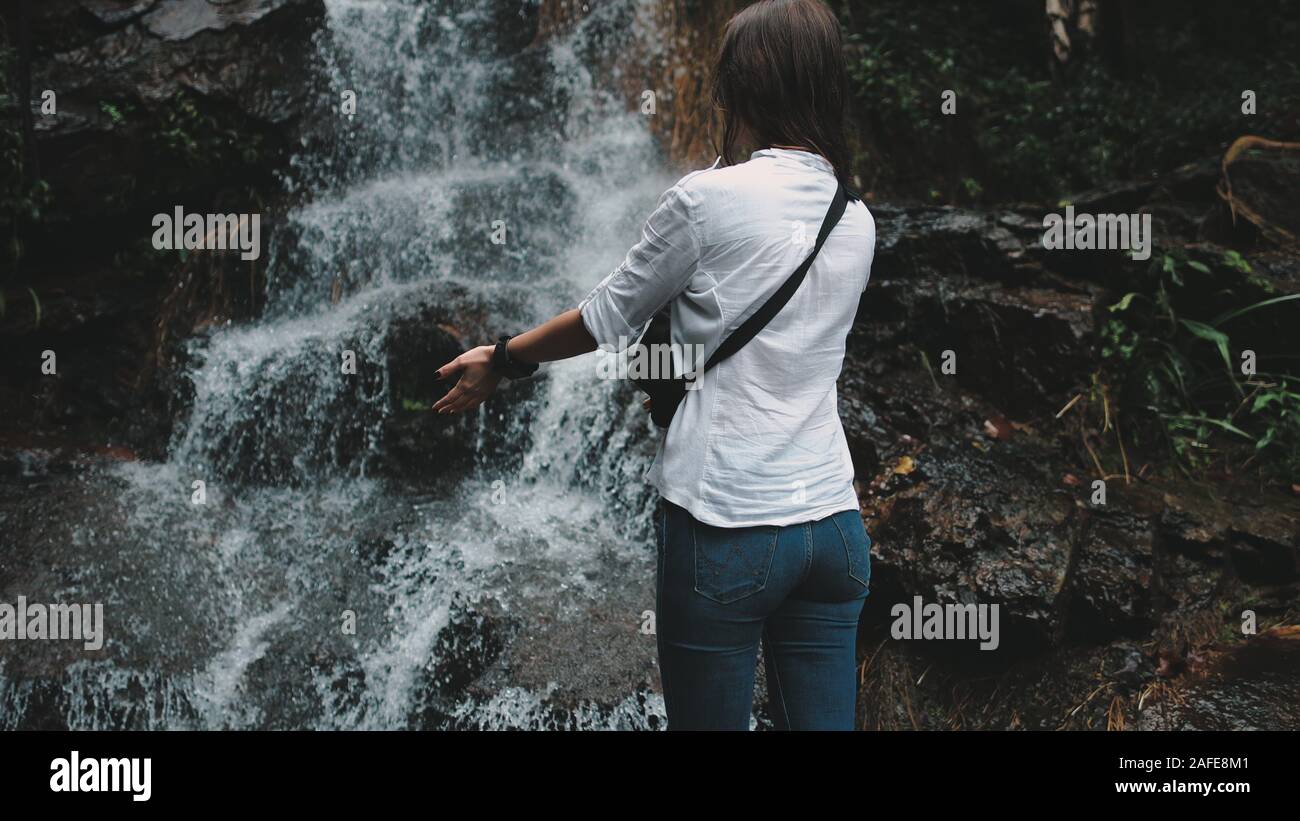 Woman Tourist Reaching out Hands to Splashing Waterfall. Caucasian Young Girl Wearing Jeans and Shirt Hike at Beautiful Canyon. Traveler Enjoy Cool Water Flood. Tropical Vacation. Stock Photo