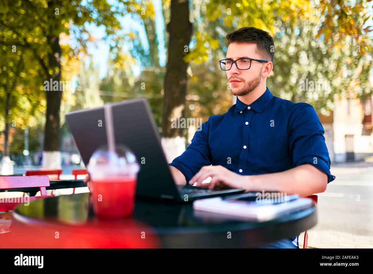 Young attractive hipster freelancer working remotely on laptop at outdoor cafe. Businessman browsing internet at coffeshop. Blogger posts to social Stock Photo