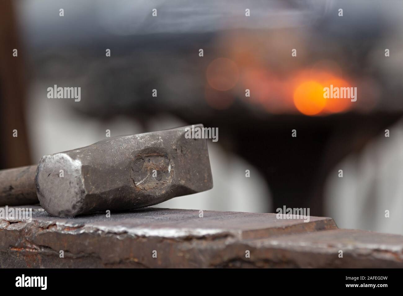 Old rusty hammer lies on the anvil with flame of brazier forge on background. Blacksmith, metalsmith, farrier tools. Close view. Stock Photo