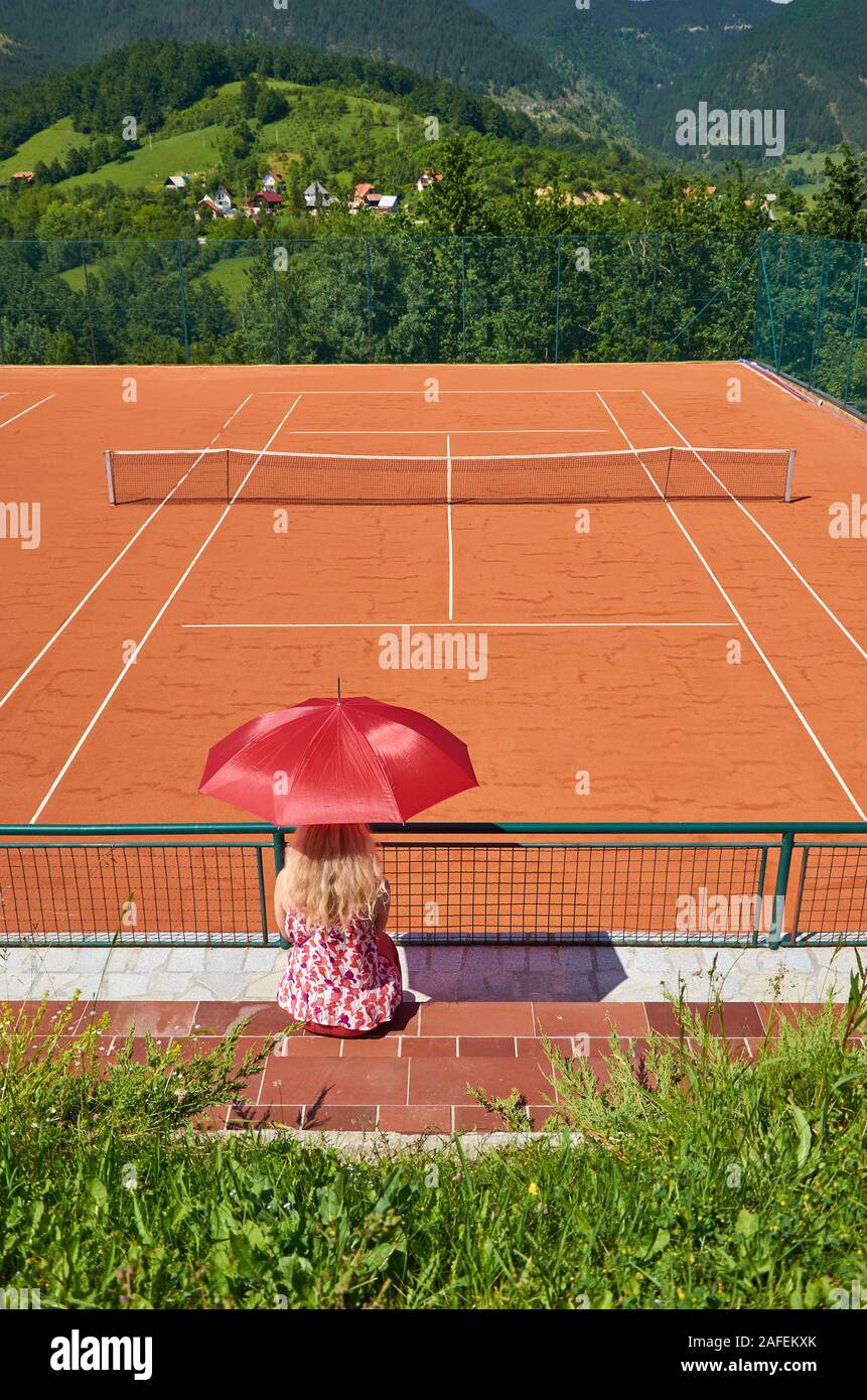 Woman with an umbrella for sun protection watching an empty tennis