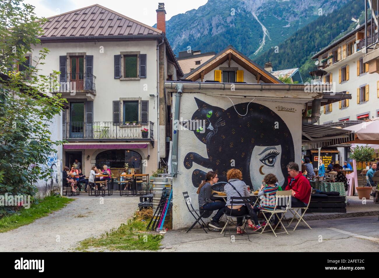 A family eats dinner at a table laid outside a Chamonix restaurant with a painting on the wall. Stock Photo