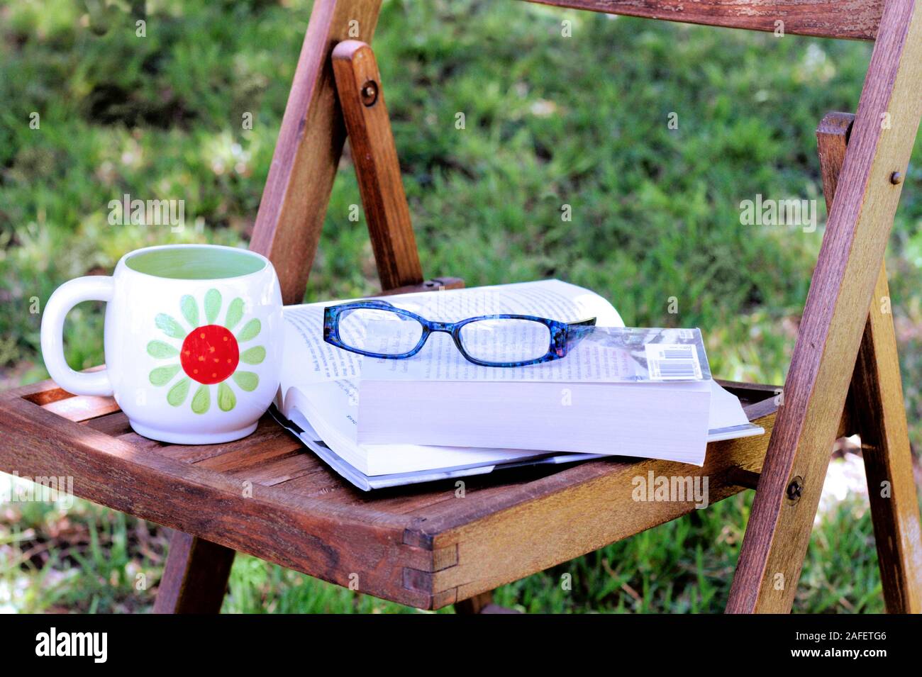 Books rest on wooden slat chair in dappled shade with reading glasses and coffee mug. Stock Photo