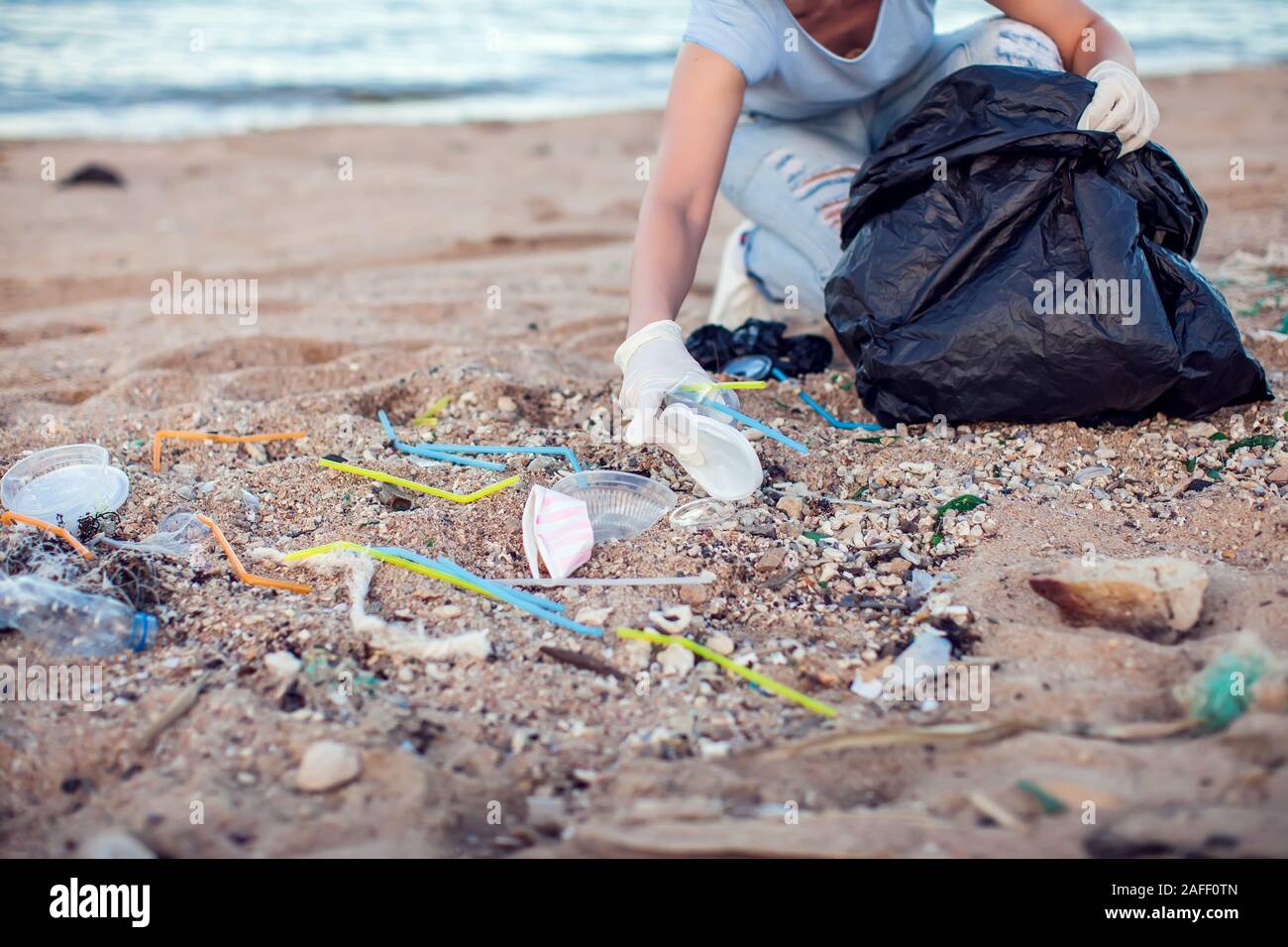 Woman in blue shirt with white gloves and big black package collecting garbage on the beach. Environmental protection and planet pollution concept Stock Photo