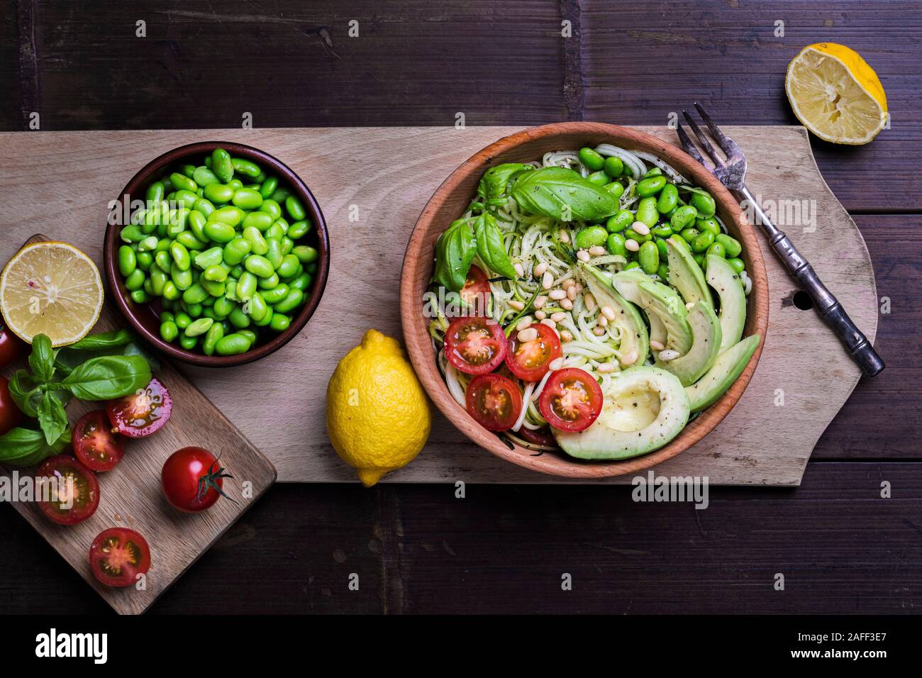 A fresh, healthy salad with zoodles zucchini noodles, baby tomatoes, avocado and edamame beans. The salad is in a wooden bowl on a dark bamboo table o Stock Photo