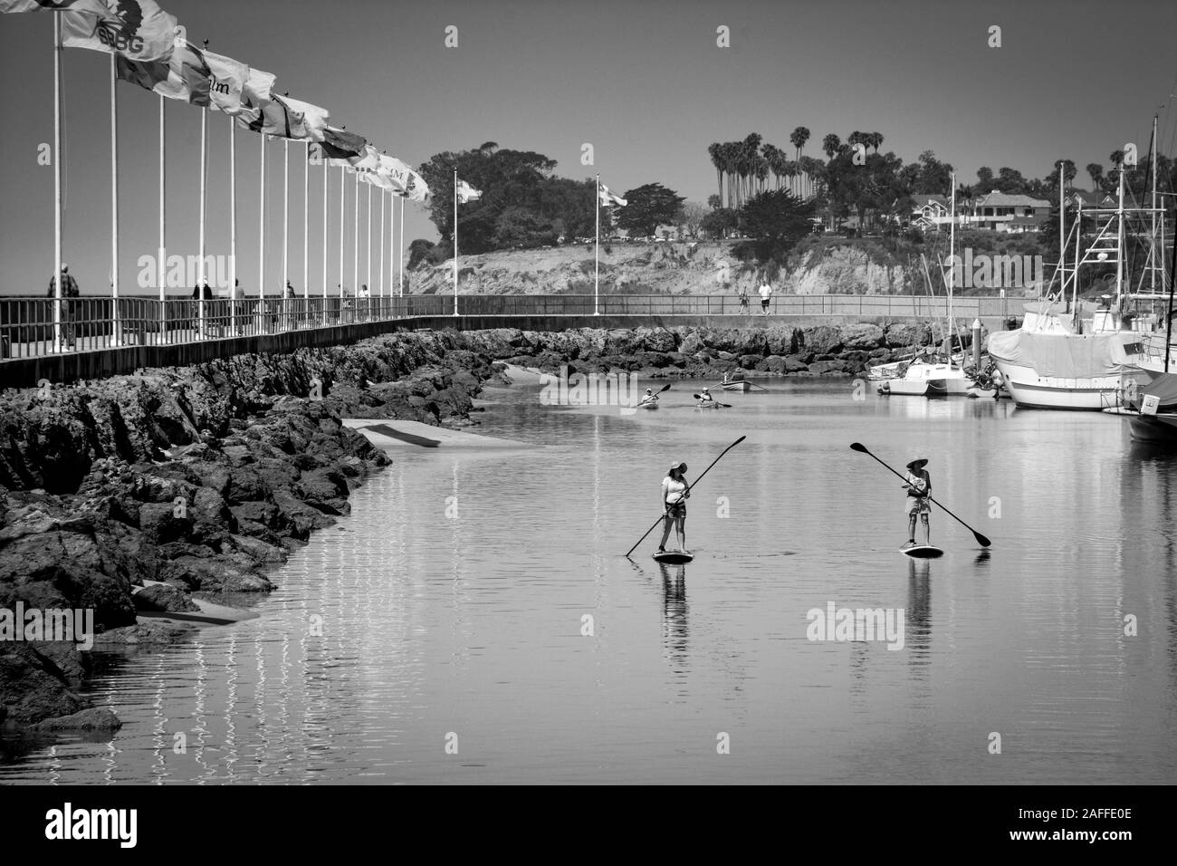Two women maneuver their stand up paddle boards through a channel near the breakwater path and marina in the Santa Barbara Harbor, Santa Barbara, CA, Stock Photo