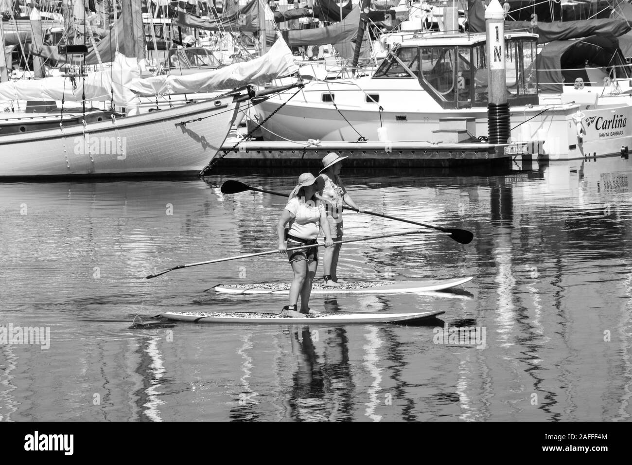 Two women maneuver their stand up paddle boards through a channel near docked boats through the marina in the Santa Barbara Harbor, Santa Barbara, CA Stock Photo