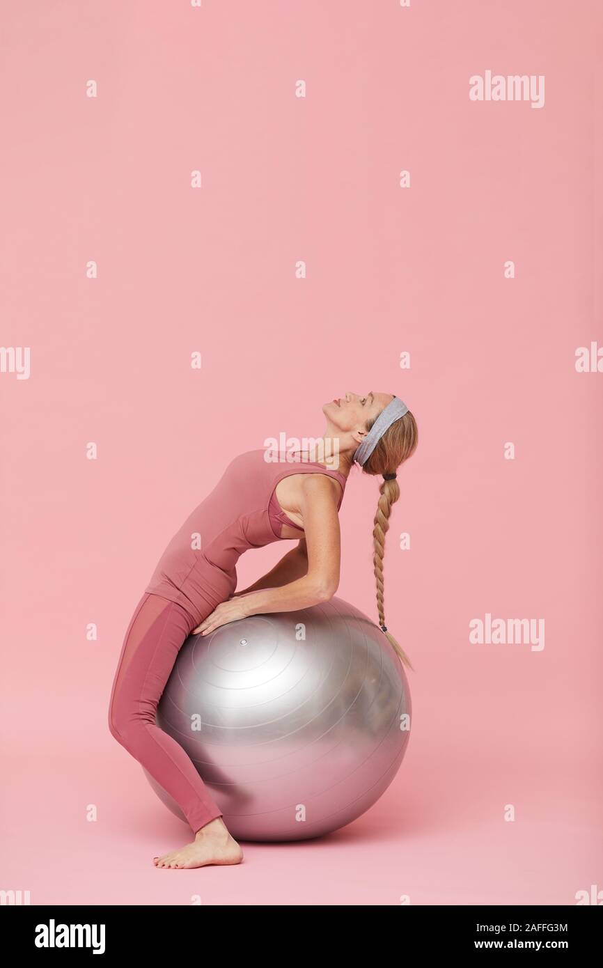 Side view portrait of active mature woman doing stretching exercises with fitness ball against pastel pink background, copy space Stock Photo