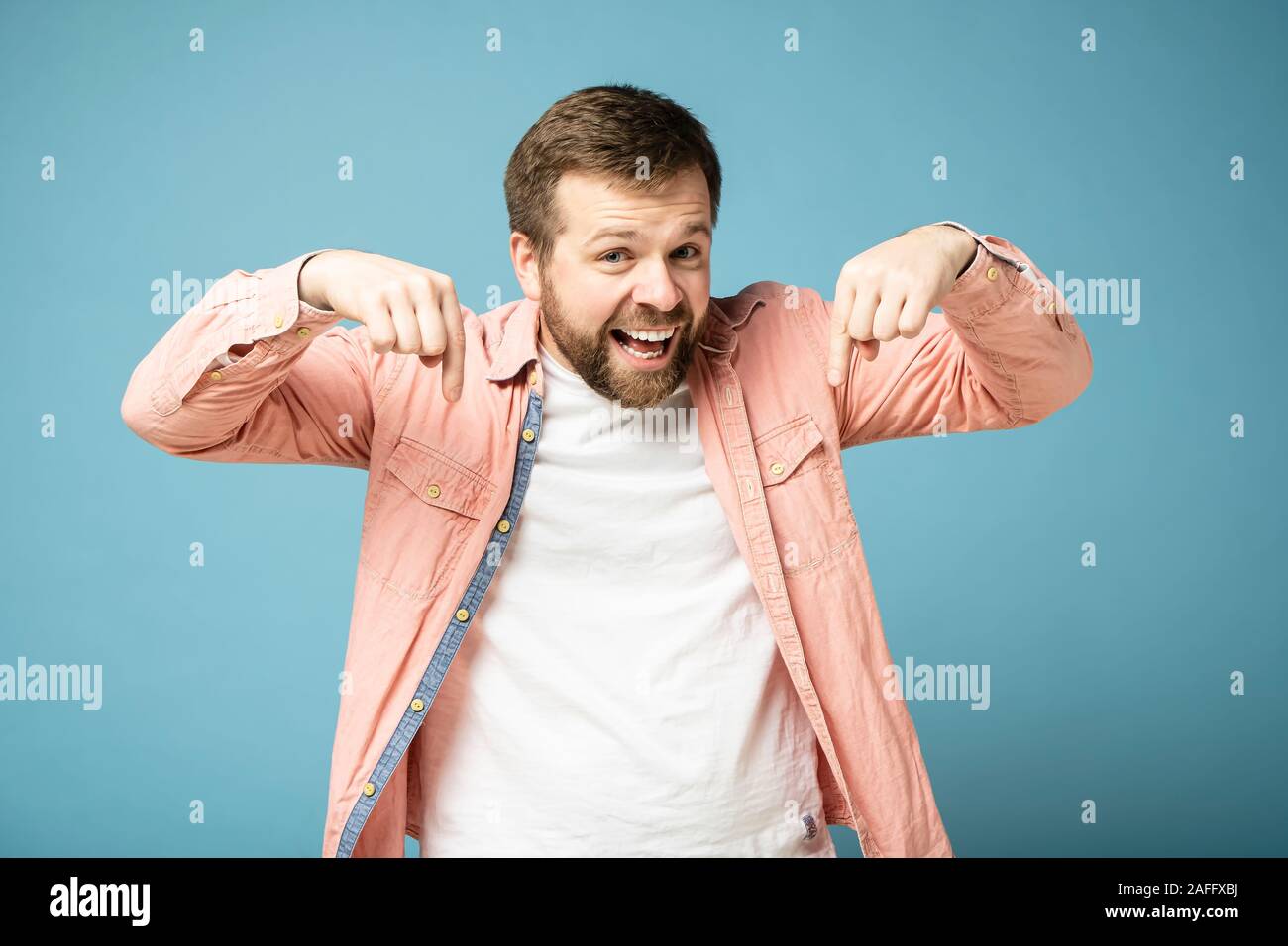 Happy bearded man looks and points his index finger down, smiling broadly and looking at the camera. Isolated on a blue background Stock Photo