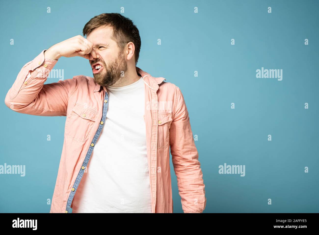Bearded man squeezes nose with fingers because of an unpleasant odor. Isolated on a blue background. Stock Photo