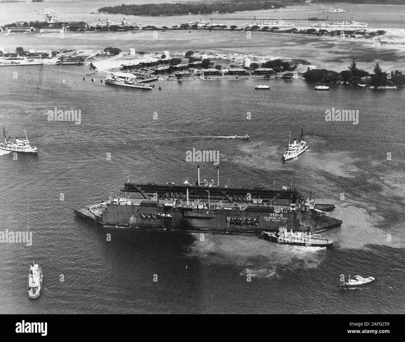 Floating Drydock YFD-2 -  Arrives at Pearl Harbor, Hawaii, on 23 August 1940, after being towed from New Orleans, Louisiana. It is still marked U.S. Naval Station New Orleans, La. USS Osceola (YT-129) is in the right foreground, assisting. One of the other tugs is probably USS Sunnadin (AT-28). An unidentified destroyer seaplane tender (AVD) is tied up at the Ford Island fuel dock, in the left center. Visible in the distance, moored on the other side of Ford Island, are (from left to right): USS Yorktown (CV-5), two destroyers, USS Wright (AV-1) and two light cruisers. Stock Photo