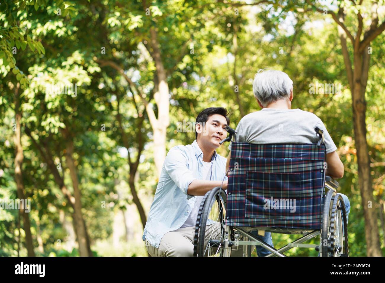 young asian adult son consoling wheelchair bound father outdoors in park Stock Photo