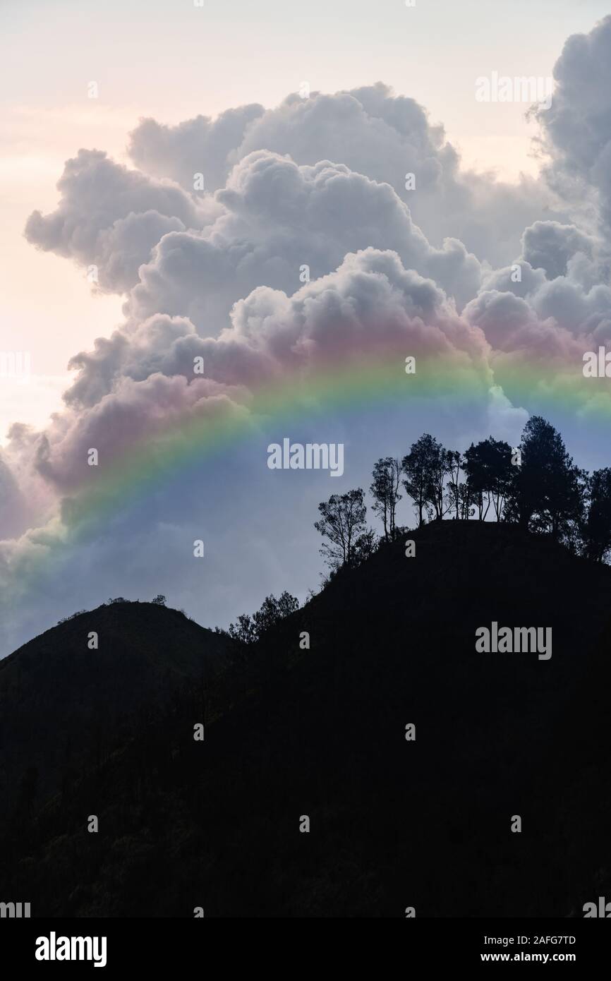 Stunning view of the silhouette of some sinuous hills with a beautiful rainbow and impressive clouds in the background. Cemoro Lawang, East Java, Indo Stock Photo